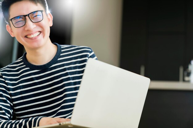 Glückliche asiatische Smart-Casual-Brille, männlich, die von zu Hause aus mit Laptop-Technologie arbeitet, mit komfortablem und entspanntem Gesichtsausdruck, modernes Zuhause mit leerem Raum im Innenraum
