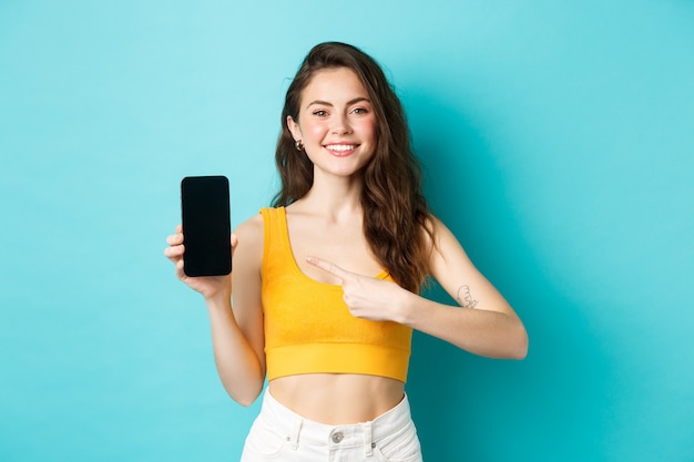 Glückliche attraktive Frau, die Werbung auf dem Smartphone-Bildschirm zeigt, auf die leere Anzeige des Telefons zeigt und lächelt, auf blauem Hintergrund stehend.