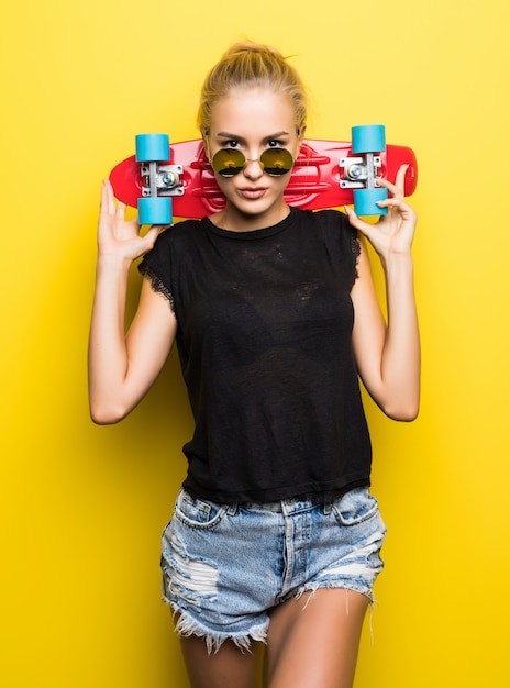 Glückliche attraktive junge Frau in Sonnenbrille, die auf Skateboard über gelbem Hintergrund sitzt