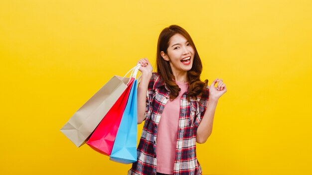 Glückliche aufgeregte junge asiatische Dame, die Einkaufstaschen mit der Hand trägt, die in der Freizeitkleidung anhebt und Kamera über gelber Wand betrachtet. Gesichtsausdruck, saisonaler Verkauf und Konsumkonzept.