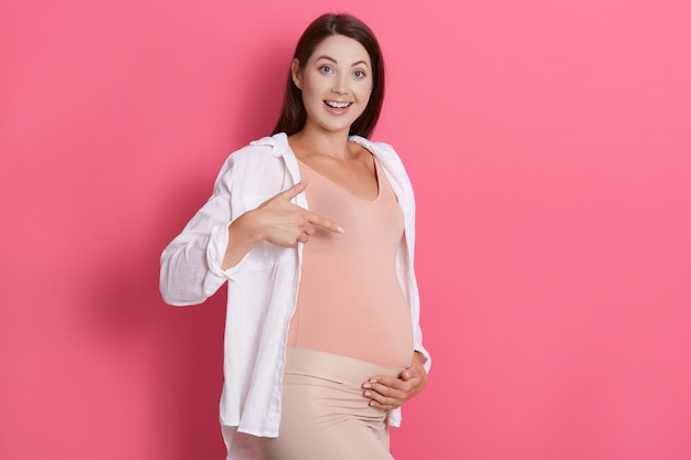 Glückliche aufgeregte schwangere Frau, die mit charmantem Lächeln auf ihren Bauch zeigt, lacht, während sie direkt in die Kamera schaut, stilvolle Kleidung trägt, hat dunkles Haar.