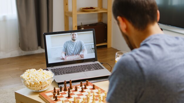 Glückliche beste Freunde, die während der Coronavirus-Quarantäne Schach per Videoanruf spielen. Bier trinken und Popcorn essen.