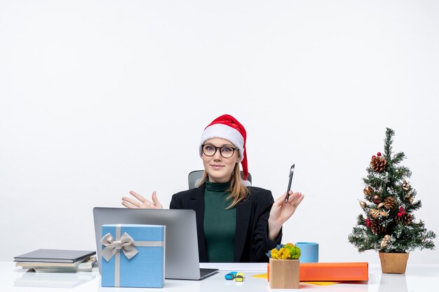 Glückliche bestimmende blonde Frau mit einem Weihnachtsmannhut, der an einem Tisch mit einem Weihnachtsbaum und einem Geschenk darauf im Büro auf weißem Hintergrund sitzt