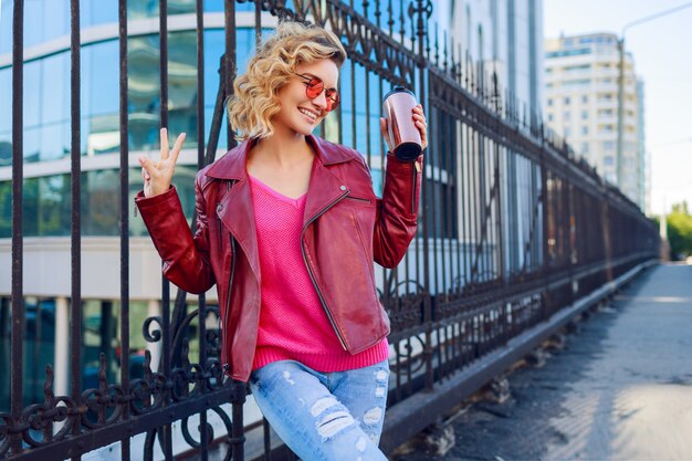 Glückliche blonde Frau, die auf modernen Straßen aufwirft, Kaffee oder Cappuccino trinkend. Stilvolles Herbstoutfit, Lederjacke und Strickpullover. Rosa Sonnenbrille.