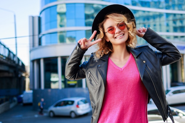 Glückliche blonde Frau, die auf modernen Straßen aufwirft. Stilvolles Herbstoutfit, Lederjacke und Strickpullover. Rosa Sonnenbrille.