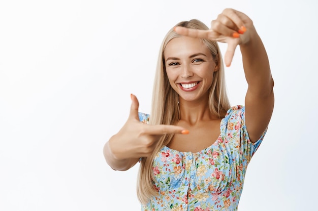 Glückliche blonde Frau, die durch Handrahmen schaut und lächelt, wenn sie etwas fotografiert, während sie kreativ vor weißem Hintergrund steht