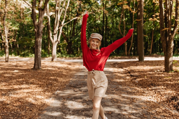 Glückliche blonde frau im schönen roten pullover und in den beigen shorts, die im herbstpark tanzen. stilvolle junge frau, die mit freude im guten weber im freien aufwirft.