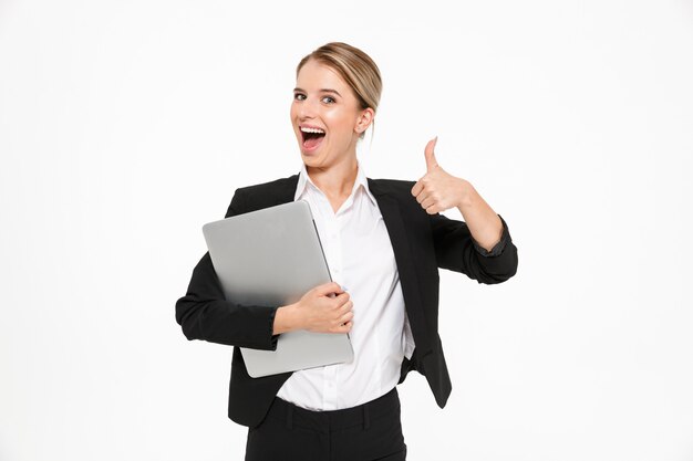 Glückliche blonde Geschäftsfrau, die Laptop-Computer hält und Daumen oben zeigt, während über weißer Wand