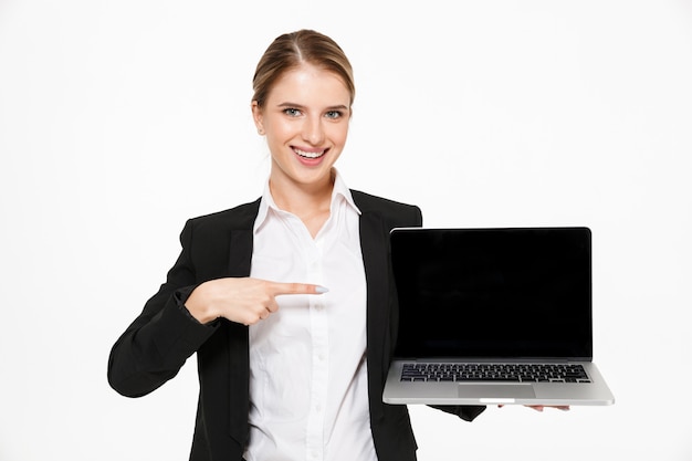 Glückliche blonde Geschäftsfrau, die leeren Laptop-Computerbildschirm zeigt und auf ihn zeigt, während über weißer Wand