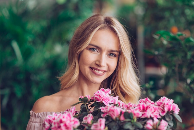 Glückliche blonde junge Frau, die hinter den rosa Blumen mit unscharfem Hintergrund steht