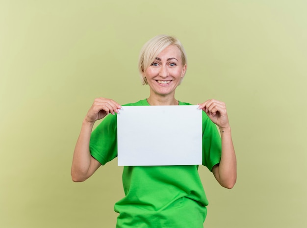 Glückliche blonde slawische Frau mittleren Alters, die leeres Papier lokalisiert auf olivgrüner Wand hält