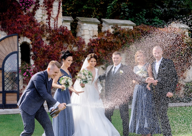 Glückliche Brautjungfern, beste Männer und Hochzeitspaar feiern Hochzeitstag im Freien mit dem Ausgießen von Champagner