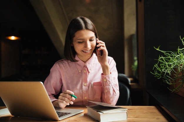 Glückliche brünette Frau, die durch den Tisch mit Laptop sitzt