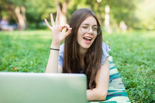 Glückliche brünette Frau in den Brillen, die auf Gras im Park mit Laptop-Computer liegen und ok Zeichen zeigen