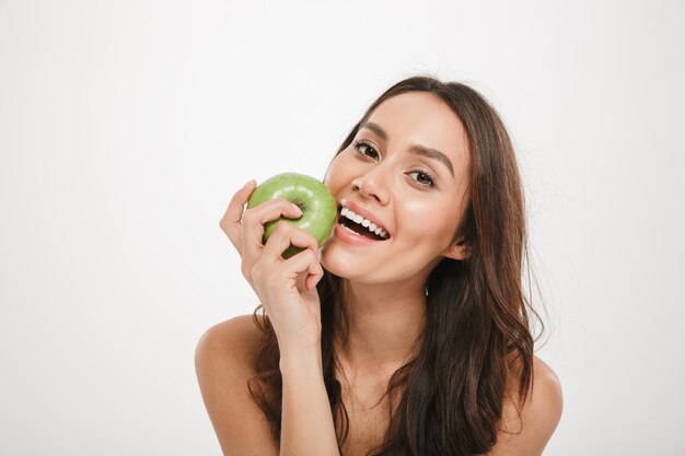 Glückliche Brunettefrau, die Apfel isst und die Kamera über Grau betrachtet