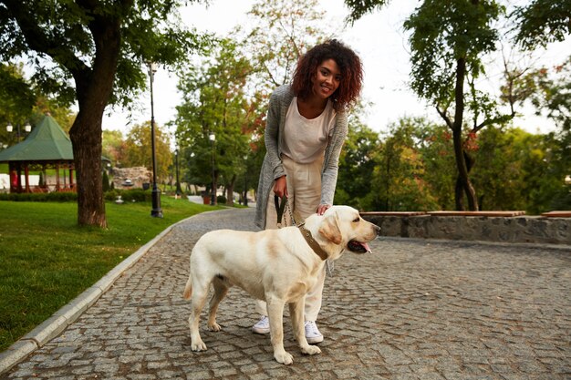 Glückliche Dame, die ihren weißen freundlichen Hund beim Gehen im Park umarmt