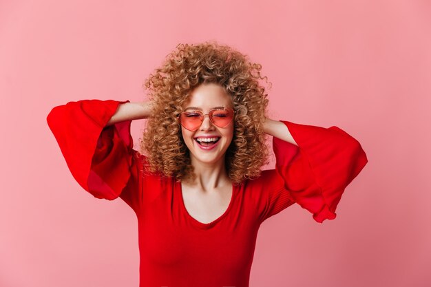 Glückliche Dame kräuselt ihre lockigen blonden Haare. Mädchen in der Brille und in der hellen Bluse lacht auf rosa Raum.