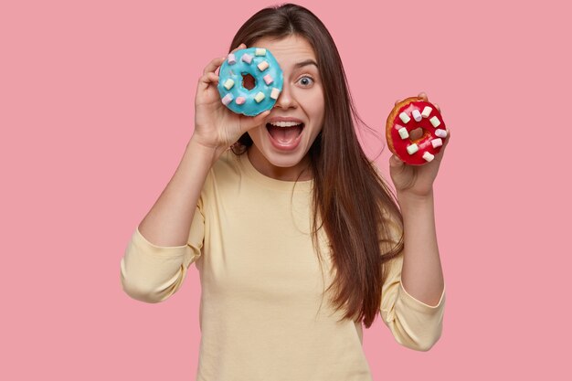 Glückliche dunkelhaarige Dame hält zwei leckere Donuts, ruft glücklich aus, trägt gelben Pullover