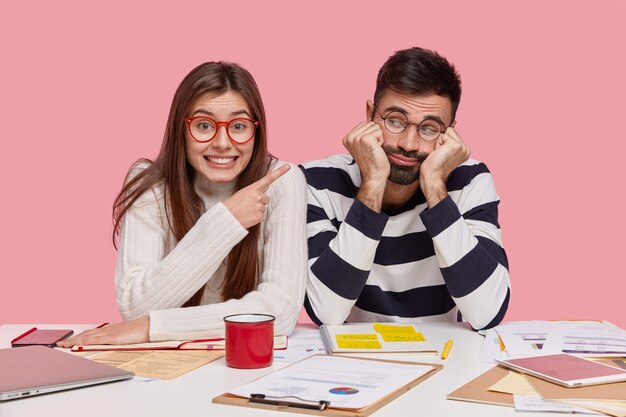 Glückliche dunkelhaarige junge Frau trägt transparente Brille, zeigt auf traurigen unrasierten Mann, trifft sich zum Vorbereiten der Hausaufgabe, posiert am Arbeitsplatz