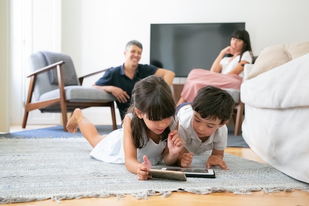 Glückliche Eltern beobachten kleine Kinder, die auf Boden im Wohnzimmer liegen und digitale Geräte zusammen verwenden.