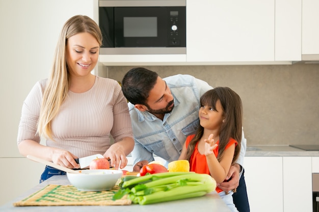 Glückliche Eltern und Kind kochen zusammen. Mädchen, das mit Papa plaudert und umarmt, während Mutter frisches Gemüse und Obst schneidet. Familienkochen oder Lifestyle-Konzept