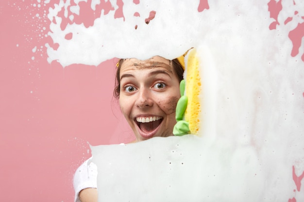 Glückliche emotionale junge Frau mit Schmutz auf ihrem Gesicht, der in der Aufregung während der Hausarbeit schaut, Räume in ihrer Wohnung putzt, Fenster wäscht, chemische Produkte und Lappen verwendet