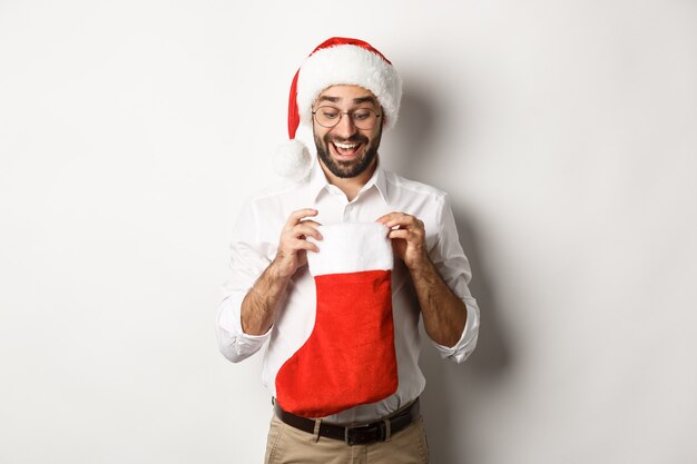 Glückliche erwachsene Mann öffnen Weihnachtssocke und schauen nach innen, Weihnachtsgeschenke für Winterferien empfangend, in der Weihnachtsmütze stehend