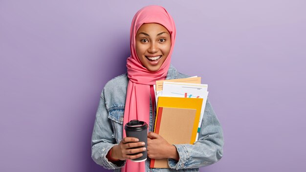 Glückliche ethnische Studentin hat Kaffeepause, hält Tasse zum Mitnehmen, trägt Notizbuch und Papiere, hat rosa Schal auf dem Kopf, islamische religiöse Ansichten, posiert drinnen. Menschen, Kultur, Tradition