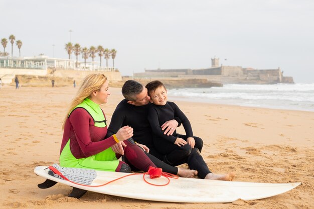 Glückliche Familie, die auf Sand nahe Surfbrett sitzt