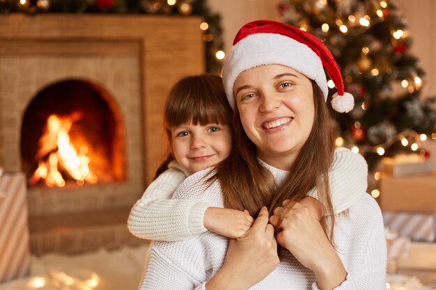 Glückliche Familie, die Zeit miteinander verbringt, Mutter und ihre kleine Tochter umarmen sich beim Sitzen auf dem Boden im festlichen Wohnzimmer mit Kamin und Weihnachtsbaum.