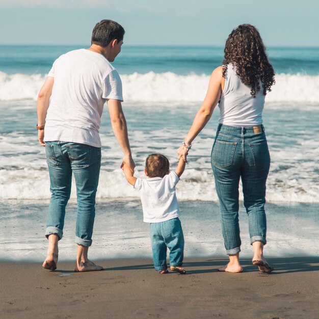 Glückliche Familie mit dem Baby, das auf Strand geht und auf Meer schaut