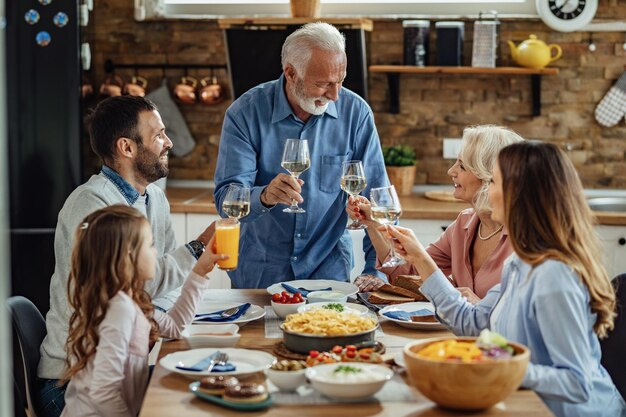 Glückliche Familie mit mehreren Generationen, die beim gemeinsamen Mittagessen am Esstisch anstößt.