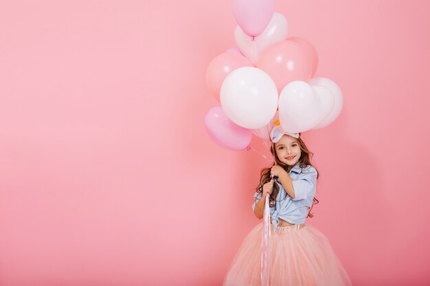 Glückliche Feier der Geburtstagsfeier mit fliegenden Luftballons des charmanten niedlichen kleinen Mädchens im Tüllrock lächelnd zur Kamera lokalisiert auf rosa Hintergrund. Charmantes Lächeln, das Glück ausdrückt. Platz für Text