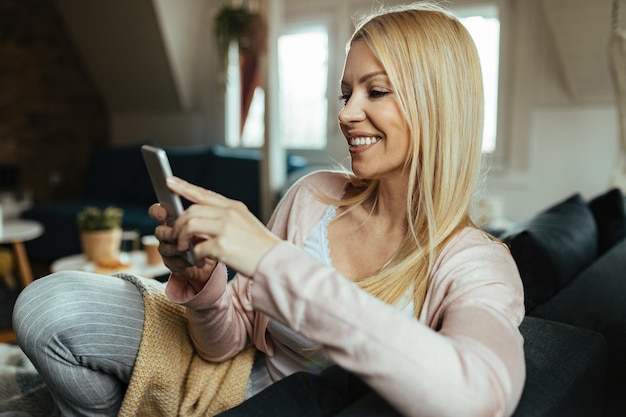 Glückliche frau, die auf dem smartphone sms schreibt, während sie sich im wohnzimmer entspannt