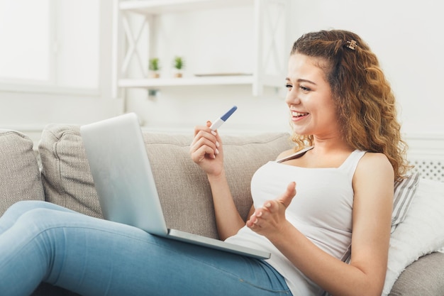 Glückliche frau, die auf laptop mit schwangerschaftstest surft, nach informationen über gesundheit und körper sucht, zu hause auf der couch liegt, platz kopiert