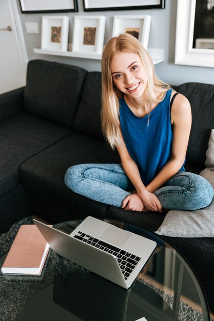 Glückliche Frau, die auf Sofa sitzt und Laptop zu Hause benutzt