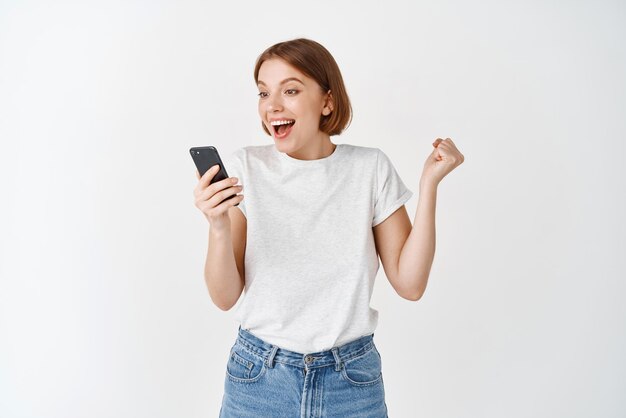 Glückliche Frau, die beim Smartphone-Videospiel gewinnt, hebt die Hand und jubelt, indem sie vor Freude ja schreit, um das Online-Ziel zu erreichen, das auf weißem Hintergrund steht