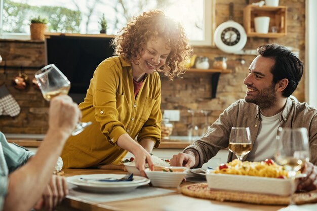 Glückliche Frau, die Essen am Tisch bringt und sich während der Mittagspause zu Hause mit ihren Freunden amüsiert