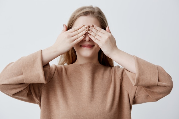 Glückliche Frau, die ihre Augen mit den Händen schließt, die Überraschung sehen, die von ihrem Freund vorbereitet wird, der steht, lächelt und auf ein Geschenk wartet. Blondes Mädchen, das ihr Gesicht mit Händen bedeckt