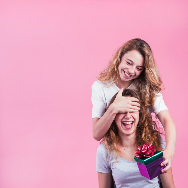 Glückliche Frau, die ihre weiblichen Augen halten Geschenkbox gegen rosa Hintergrund bedeckt