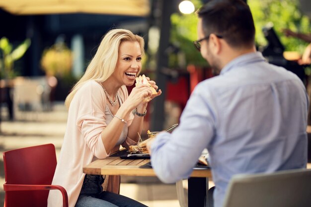 Glückliche Frau, die mit ihrem Freund spricht, während sie ein Sandwich in einem Restaurant im Freien isst