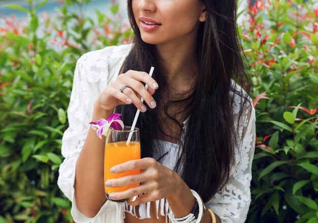 glückliche Frau, die mit leckerem frischem Orangensaft im trendigen tropischen Boho-Outfit in ihren Ferien entspannt.