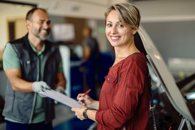 Glückliche Frau, die Papierkram in der Autowerkstatt unterschreibt und in die Kamera schaut