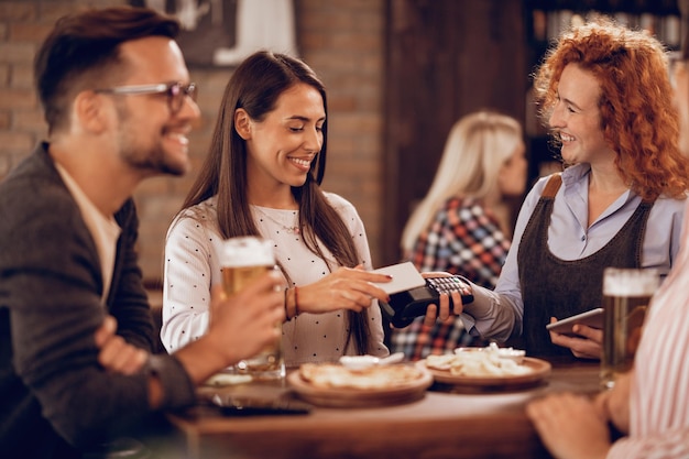 Glückliche Frau, die per kontaktloser Smartphone-Zahlung in einer Bar an eine Kellnerin zahlt