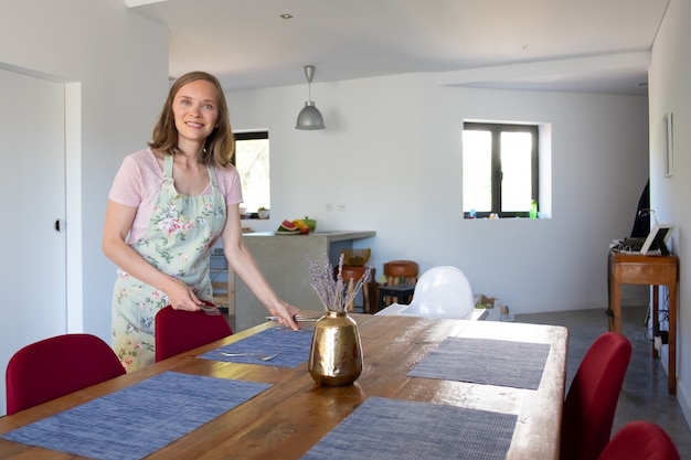 Kostenloses Foto glückliche frau, die schürze trägt und esstisch für familienessen zu hause dient. essen zu hause oder hausfrau konzept