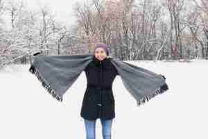 Kostenloses Foto glückliche frau, die warme kleidung trägt und den gemütlichen schal steht auf schneebedecktem land im winter hält