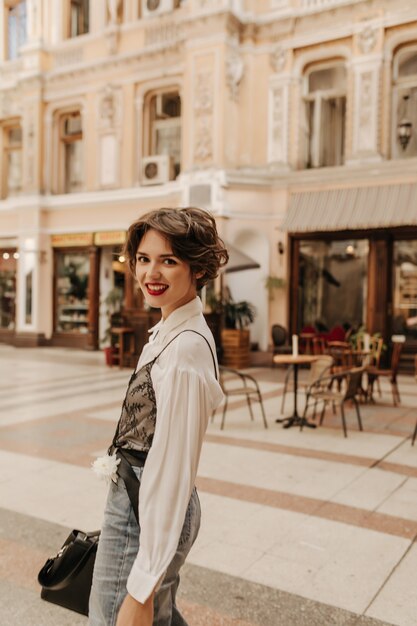 Glückliche Frau im hellen Hemd und in den Jeans in der Stadt. Moderne Frau mit kurzen Haaren und hellen Lippen, die an Straße lächeln.