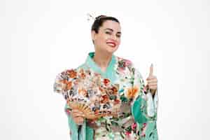 Kostenloses Foto glückliche frau im traditionellen japanischen kimono, der handfächer hält und breit lächelt, zeigt ok zeichen auf weiß
