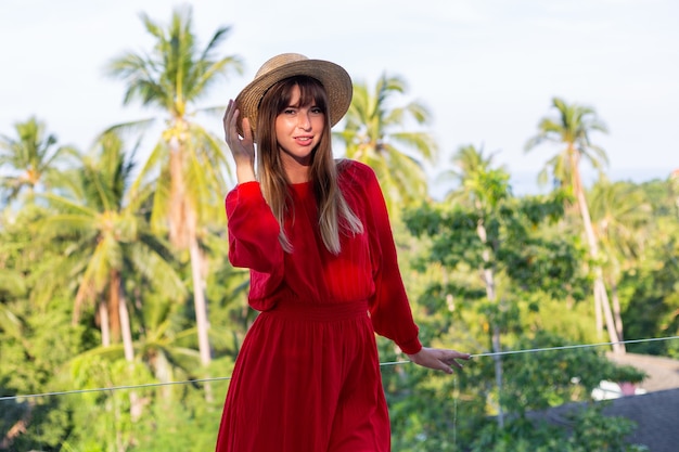 Glückliche Frau im Urlaub im roten Sommerkleid und im Strohhut auf Balkon mit tropischem Blick auf Meer und Plam-Bäume.