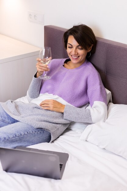 Glückliche Frau im warmen Pullover im Bett mit Glas Wein allein durch das Ausruhen beim Anschauen der Filmkomödie Lächeln lachen